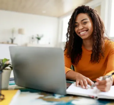 Accounting student smiling while studying financial statements on a laptop and taking notes at home